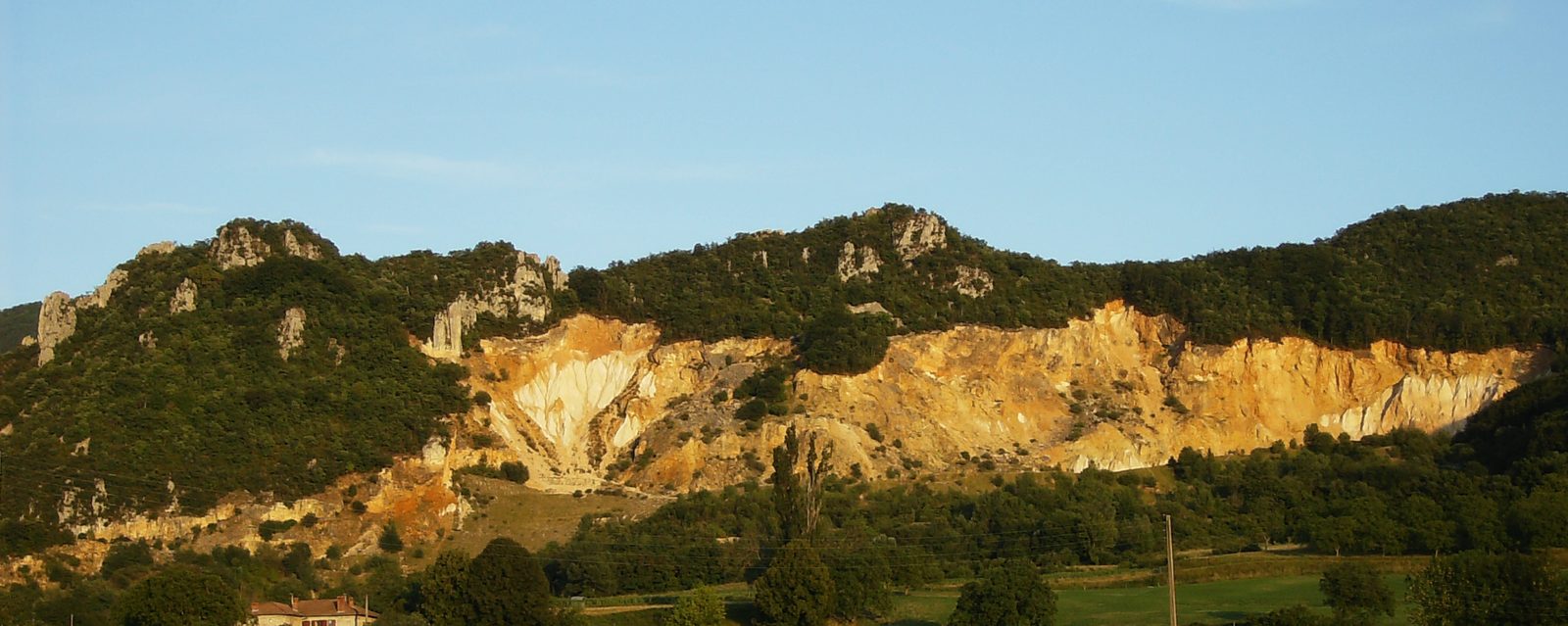 Une falaise jaune et blanche entaillée par une ancienne carrière, sous une crête boisée