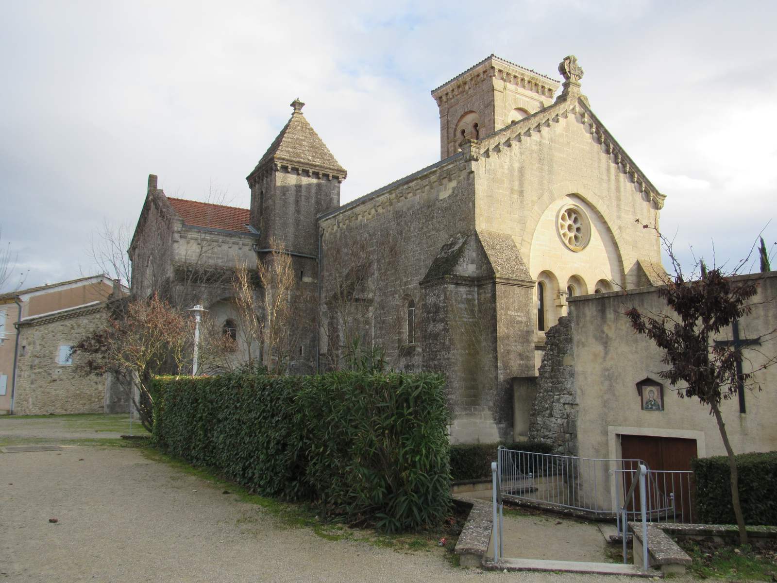 La façade de la basilique Sainte-Anne de Bonlieu, en pierre claire, avec sa rosace, son clocher carré et une tourelle à toit pointu, derrière une haie