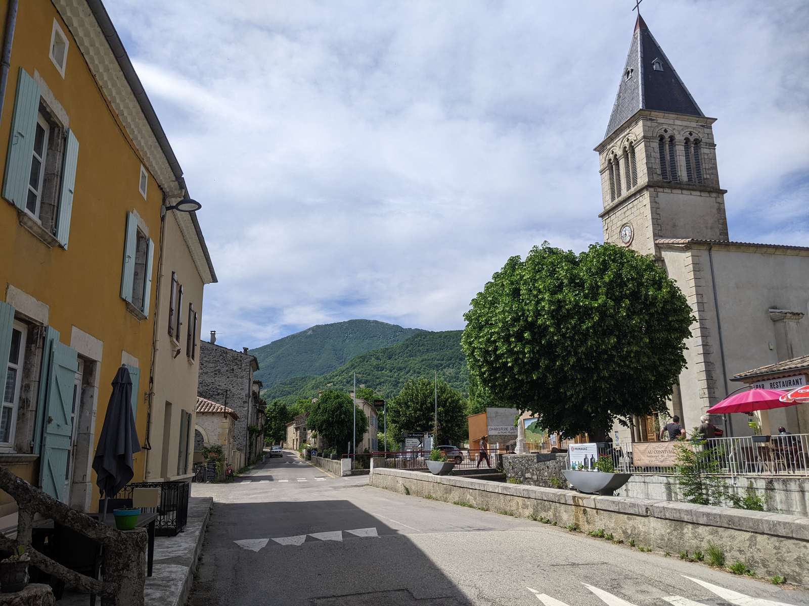 La traversée de Bouvières, l'église et son clocher pointu au bord de la route, des montagnes boisées au fond