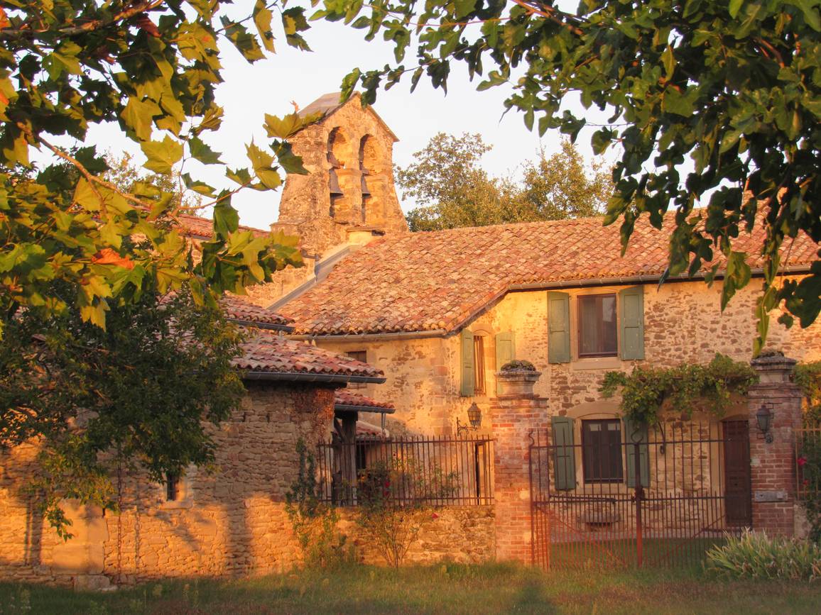 La chapelle de Gillons dans la lumière du soir, clocher-mur à deux baies au-dessus d'une maison de pierre aux volets verts, derrière une grille
