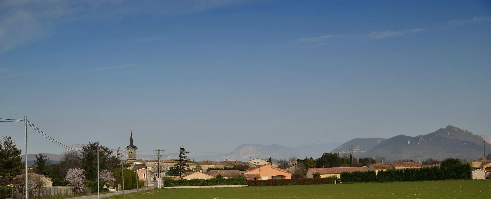 Cléon-d'Andran vu depuis un champ, le clocher pointu au-dessus des toits, avec les montagnes à l'horizon sous un ciel bleu