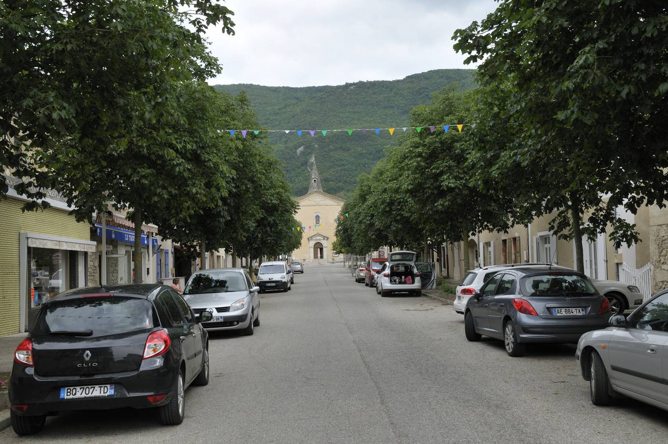 L'avenue des Marronniers à Hostun, une rue droite bordée d'arbres qui mène à une église, voitures garées de chaque côté