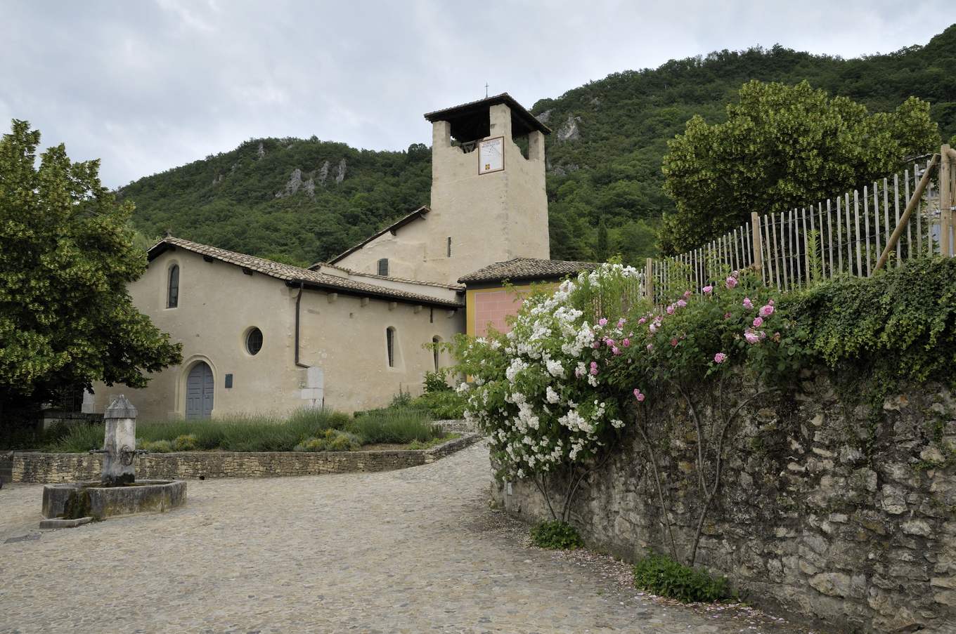 L'église Saint-Martin d'Hostun, nef claire et clocher carré, au bout d'une place pavée avec une fontaine et un mur fleuri, devant une colline boisée