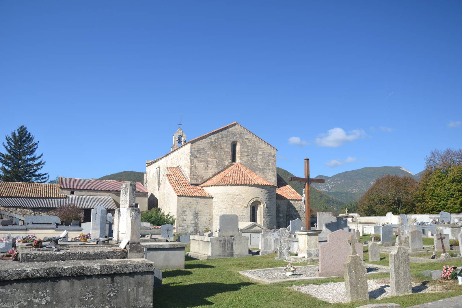 L'église romane Notre-Dame-de-Calma vue de l'est, son abside ronde et son transept couverts de tuiles, au milieu des tombes du cimetière, sous un ciel bleu