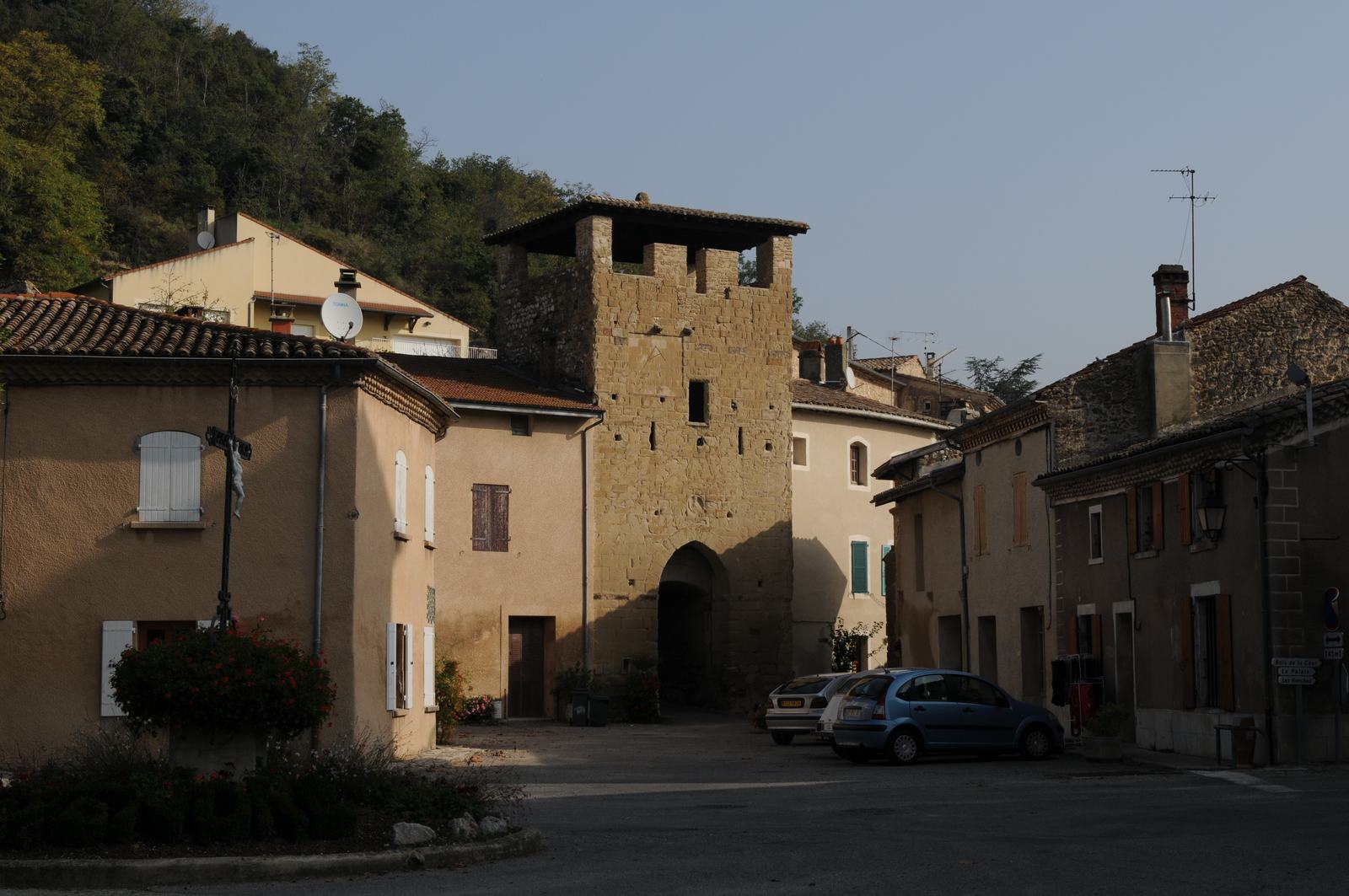 La porte fortifiée de Montvendre, haute tour carrée de pierre ocre percée d'un passage en arc, entre des maisons crépies, avec une colline boisée derrière