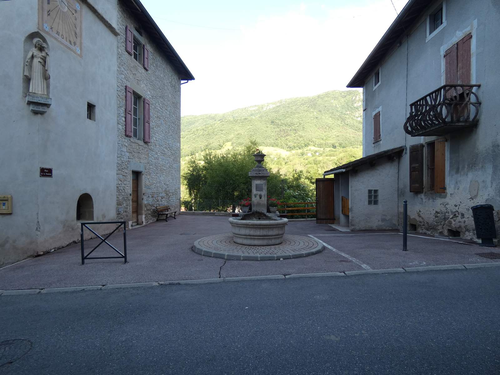 La place de la Fontaine à Oriol-en-Royans, sa fontaine de pierre, le clocher avec son cadran solaire et une statue, devant les collines boisées