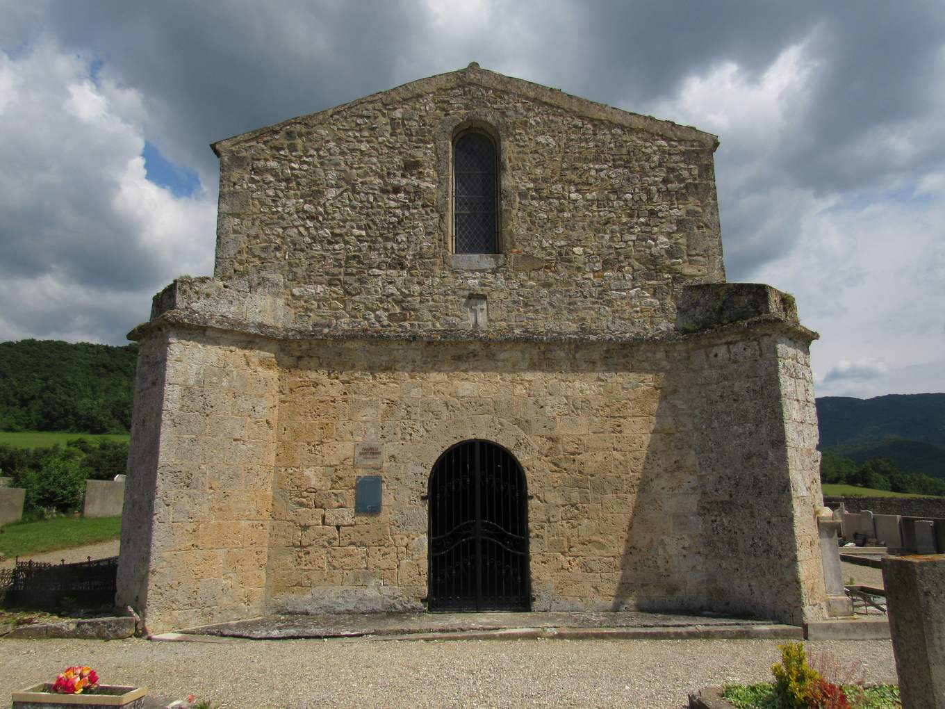La façade de la chapelle Saint-Pierre de Peyrus, haut pignon de pierre percé d'une fenêtre, et devant elle un porche massif aux contreforts obliques ouvert par une porte en plein cintre grillagée