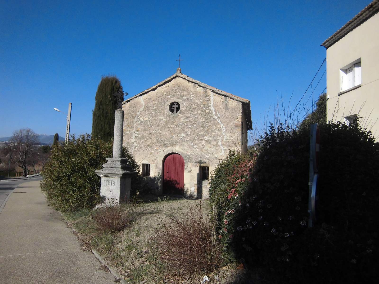 La chapelle Notre-Dame-du-Cadenet à Piégon, façade de pierre à porte rouge et oculus, une croix devant