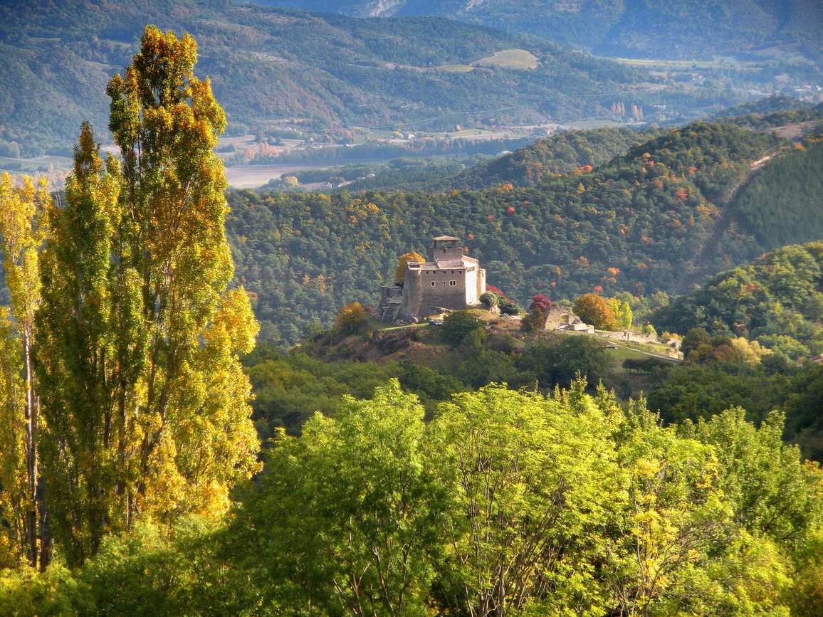 Le château de Piégros, bâtiment de pierre à tour carrée posé sur une butte boisée, entre des peupliers aux couleurs d'automne et la vallée de la Drôme au fond