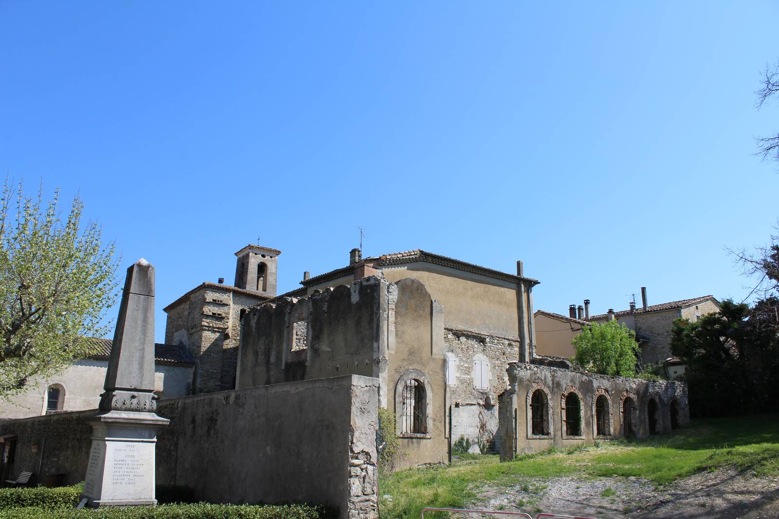 Des murs anciens à arcades en ruine à côté d'une église à clocher carré et d'un monument aux morts en obélisque, sous un ciel bleu