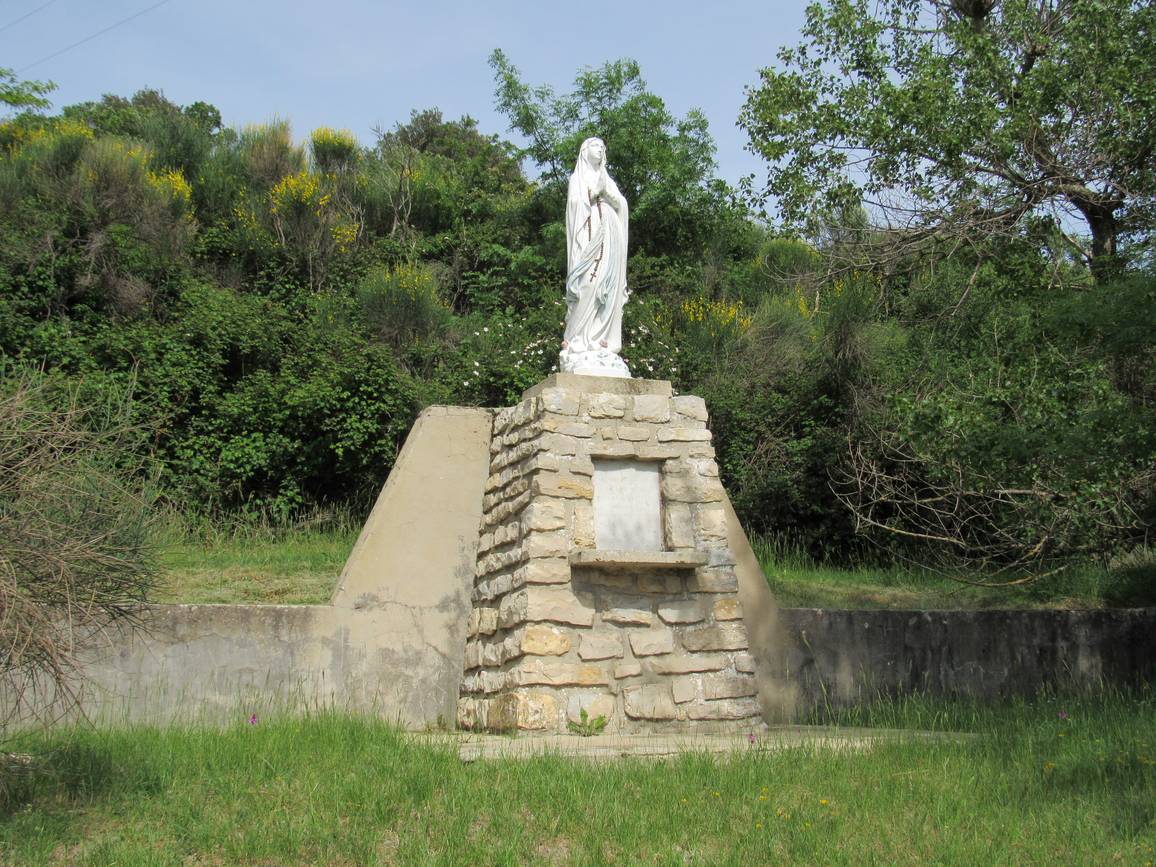 La statue blanche de la Vierge du Vœu sur un socle de pierre, au bord d'un talus fleuri de genêts