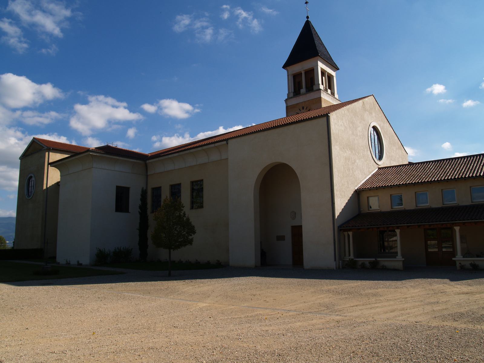 L'église abbatiale de Triors, bâtiment clair à toits de tuiles avec un grand porche en arc et un clocher carré à flèche sombre, devant une cour de gravier